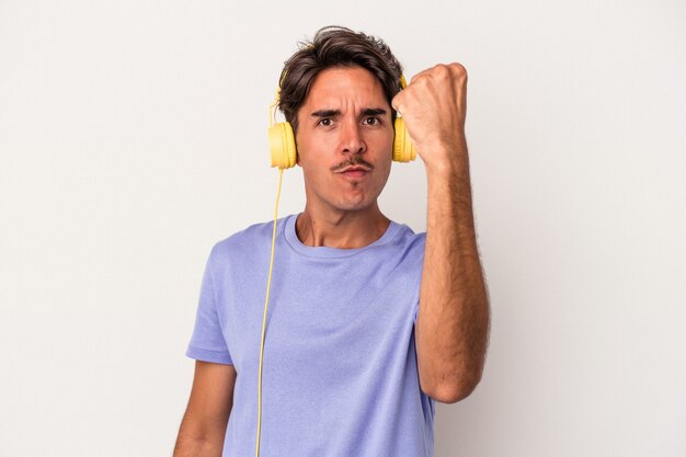 Young mixed race man listening to music isolated on blue background showing fist to camera, aggressive facial expression.