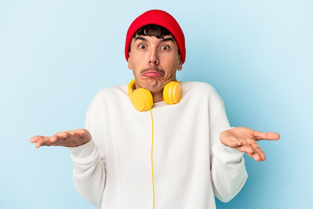 Young mixed race man listening to music isolated on blue background shrugs shoulders and open eyes confused.