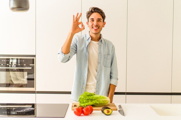 Young mixed race man preparing a salad for lunch cheerful and confident showing ok gesture.