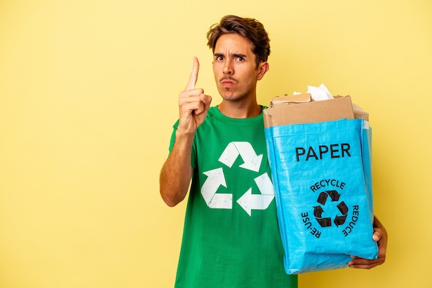 Young mixed race man recycling paper isolated on yellow background showing number one with finger.