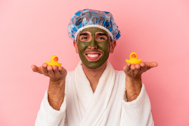 Young mixed race man wearing beauty facial mask holding rubber ducks isolated on pink background