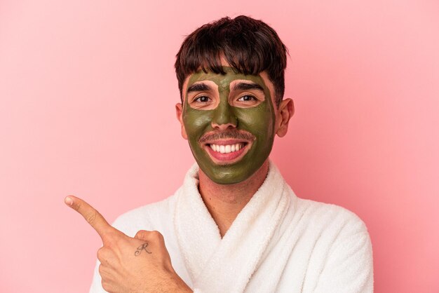 Young mixed race man wearing beauty facial mask isolated on pink background