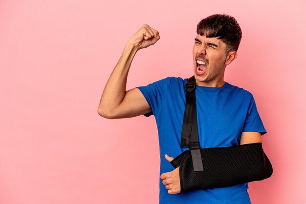 Young mixed race man with broken hand isolated on pink background raising fist after a victory, winner concept.