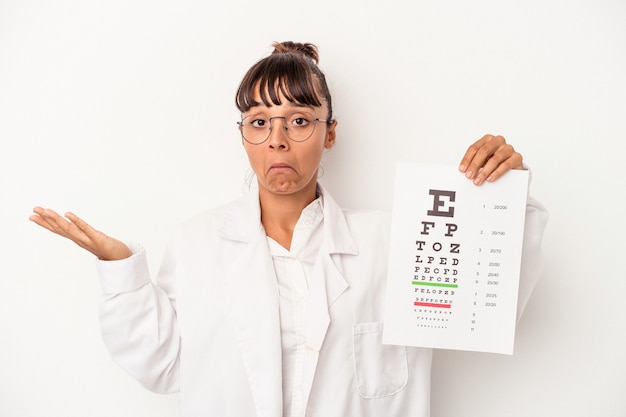 Young mixed race optician woman doing a test isolated on white background  shrugs shoulders and open eyes confused.