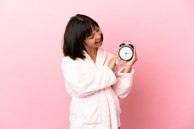 Young mixed race pregnant woman isolated on pink background in pajamas and holding clock with happy expression