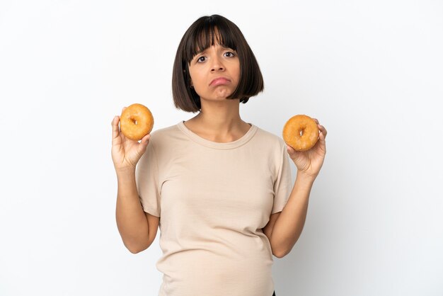슬픈 표정으로 도넛을 들고 흰 배경에 고립 된 젊은 혼혈 임신 한 여자