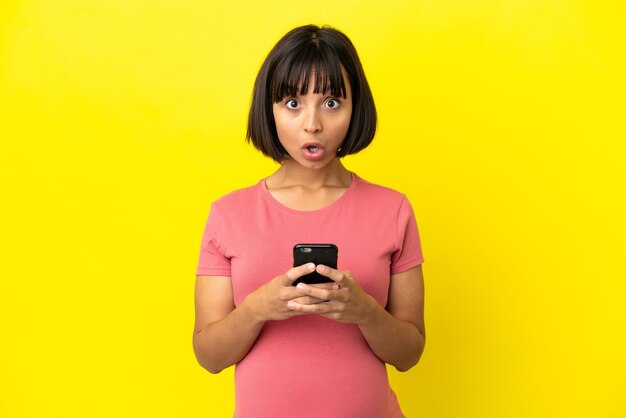 Young mixed race pregnant woman isolated on yellow background looking at the camera while using the mobile with surprised expression