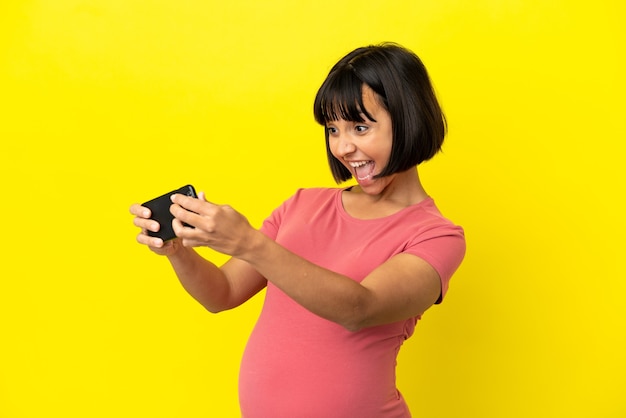 Young mixed race pregnant woman isolated on yellow background playing with the mobile phone