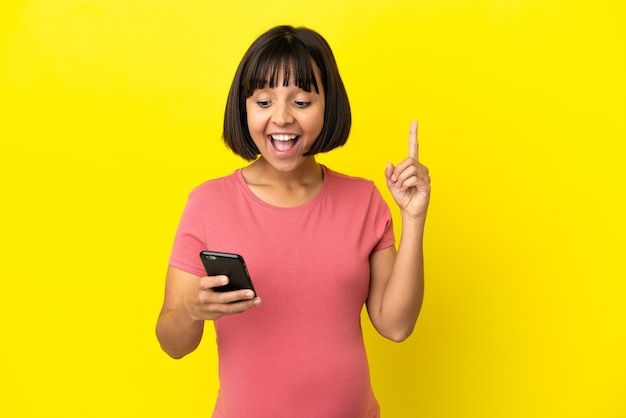 Молодая беременная женщина смешанной расы изолирована на желтом фоне, используя мобильный телефон и поднимая палец