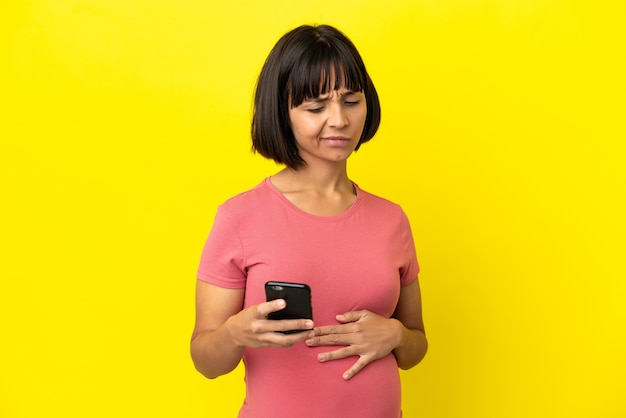 Young mixed race pregnant woman isolated on yellow background using mobile phone