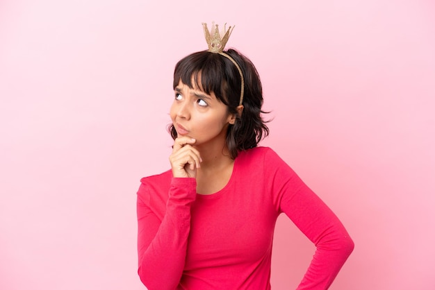 Young mixed race princess with crown isolated on pink background having doubts and with confuse face expression