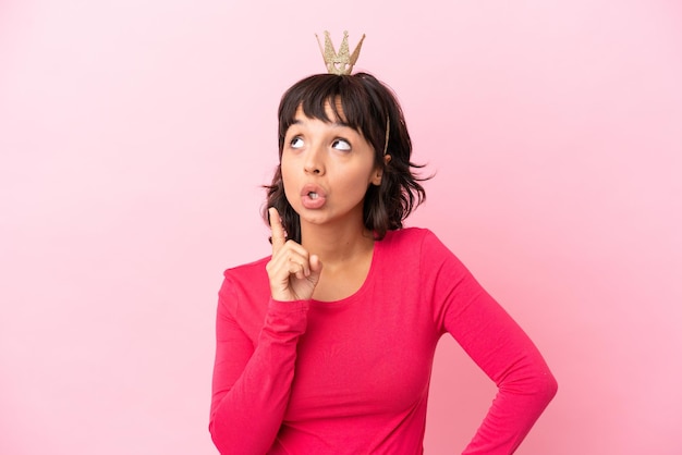 Young mixed race princess with crown isolated on pink background thinking an idea pointing the finger up
