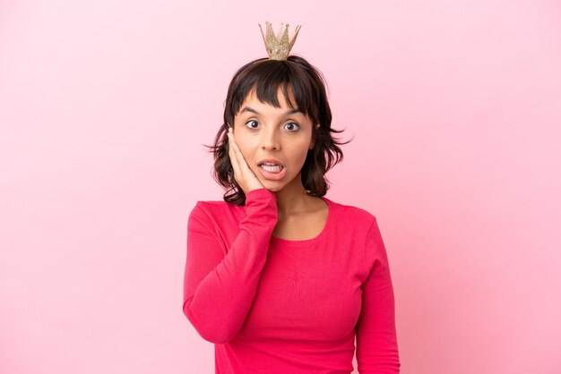 Young mixed race princess with crown isolated on pink background with surprise and shocked facial expression