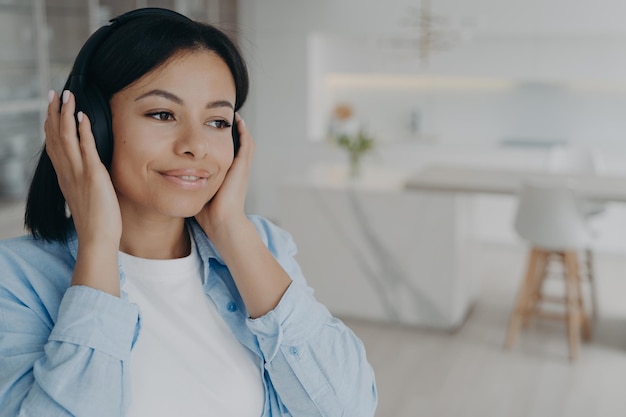 Young mixed race romantic lady is listening to radio in headphones dreaming and smiling