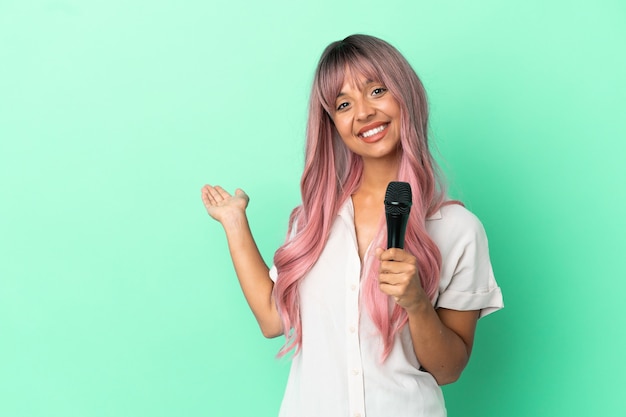 Giovane donna cantante di razza mista con i capelli rosa isolata su sfondo verde che estende le mani di lato per invitare a venire