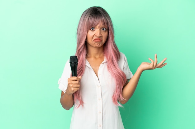 Young mixed race singer woman with pink hair isolated on green background having doubts while raising hands