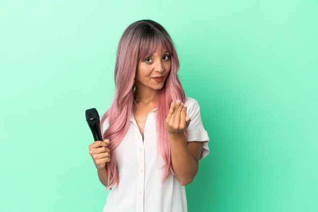 Young mixed race singer woman with pink hair isolated on green background inviting to come with hand. Happy that you came