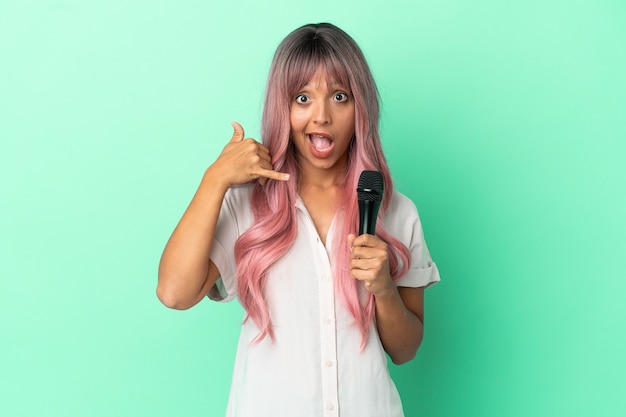 Молодая певица смешанной расы с розовыми волосами, изолированными на зеленом фоне, делая телефонный жест. Перезвони мне знак