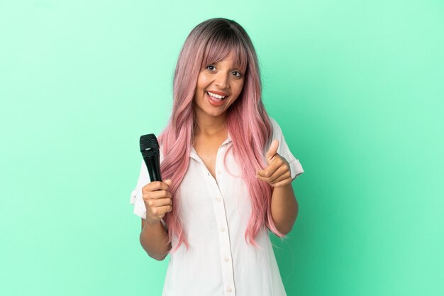 Молодая певица смешанной расы с розовыми волосами изолирована на зеленом фоне с большими пальцами руки вверх, потому что произошло что-то хорошее