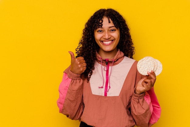 Young mixed race sport woman holding a rice cakes isolated on yellow smiling and raising thumb up