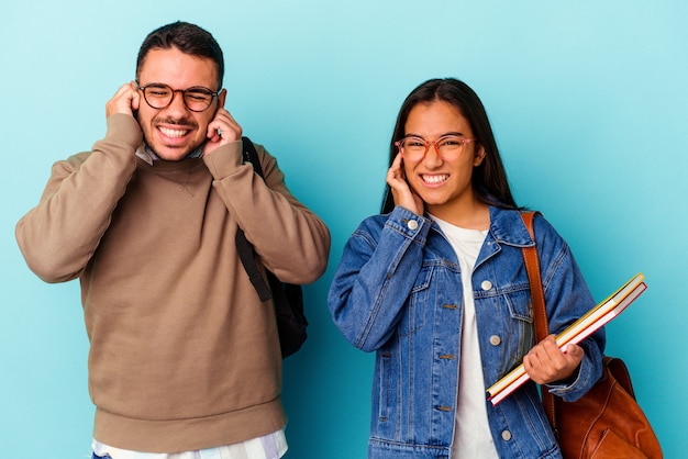 Young mixed race student couple isolated on blue background covering ears with hands.