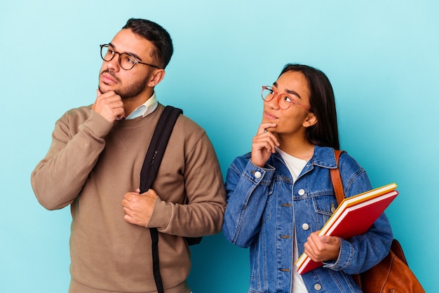 Young mixed race student couple isolated on blue background looking sideways with doubtful and skeptical expression.