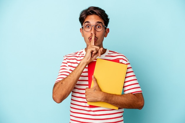 young mixed race student man holding books isolated on blue background keeping a secret or asking for silence.