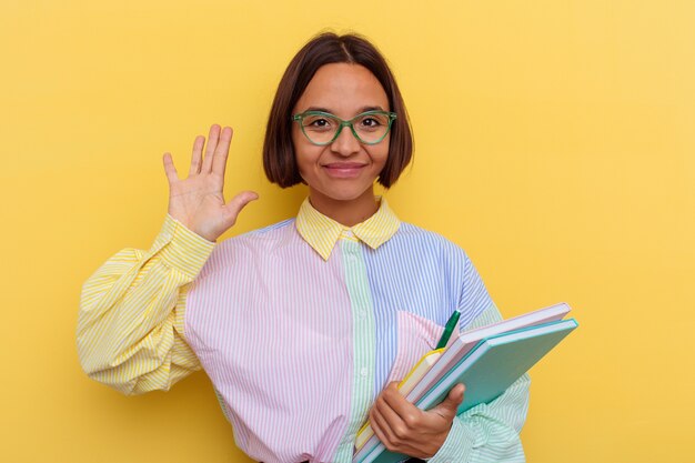 黄色の壁に孤立した若い混血の学生女性は、指で5番を示す陽気な笑顔を浮かべています。