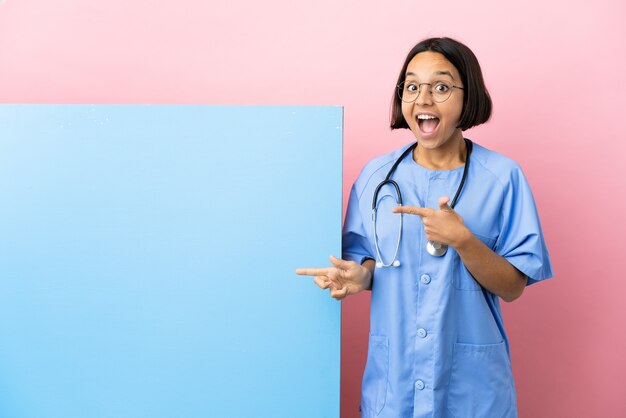 Young mixed race surgeon woman with a big banner isolated background surprised and pointing side