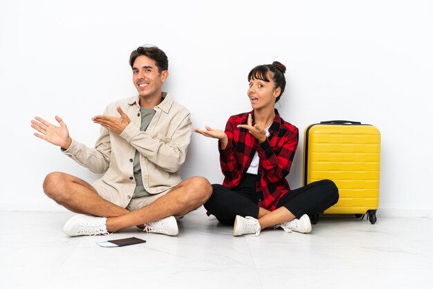 Young mixed race travelers couple sitting on the floor isolated on white background extending hands to the side for inviting to come