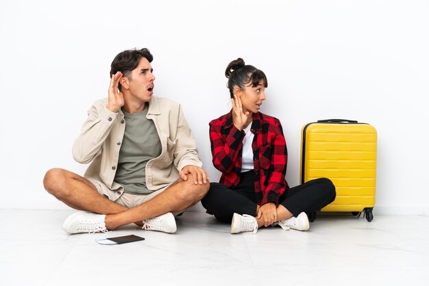 Young mixed race travelers couple sitting on the floor isolated on white background listening to something by putting hand on the ear