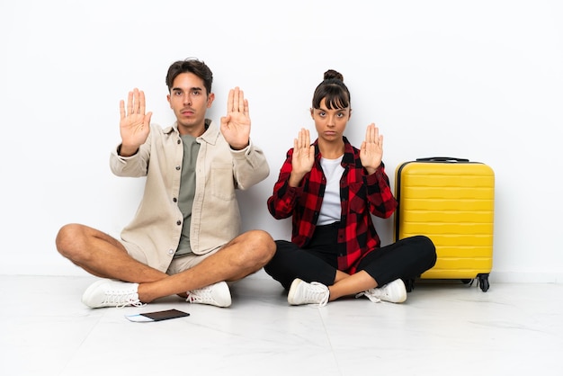 Young mixed race travelers couple sitting on the floor isolated on white background making stop gesture for disappointed with an opinion