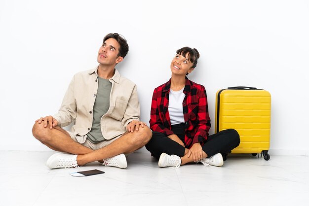 Young mixed race travelers couple sitting on the floor isolated on white background posing with arms at hip and laughing