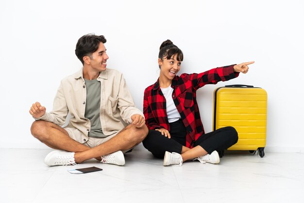 Young mixed race travelers couple sitting on the floor isolated on white background presenting an idea while looking smiling towards