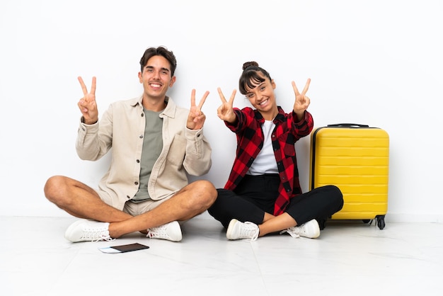 Young mixed race travelers couple sitting on the floor isolated on white background smiling and showing victory sign with both hands