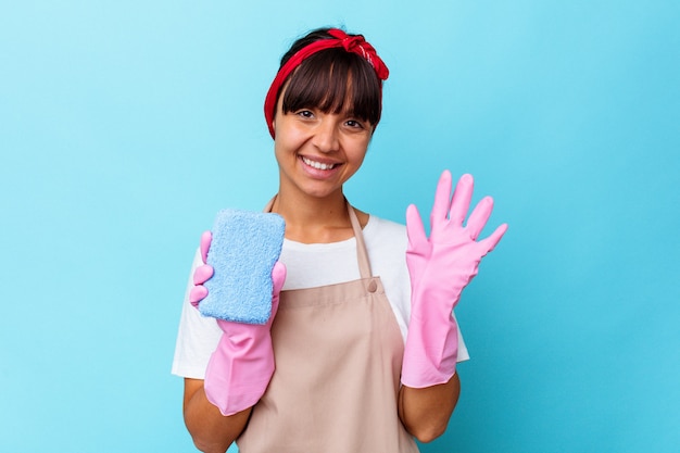 파란색 배경에 격리된 집을 청소하는 젊은 혼혈 여성이 손가락으로 5번을 보여주며 밝게 웃고 있습니다.