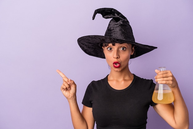 Young mixed race woman disguised as a witch holding potion isolated on purple background  pointing to the side