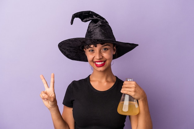 Молодая женщина смешанной расы, замаскированная как ведьма, держащая зелье, изолирована на фиолетовом фоне, показывая номер два пальцами.
