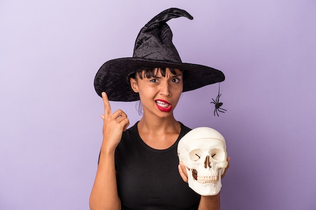 Young mixed race woman disguised as a witch holding a skull isolated on purple background  showing a disappointment gesture with forefinger.