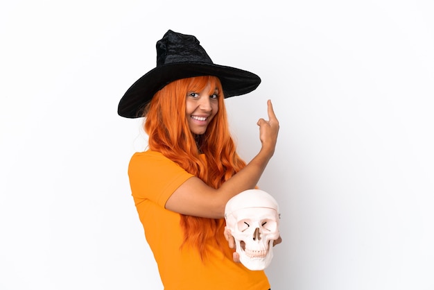 Young mixed race woman disguised as witch holding a skull\
isolated on white background pointing back