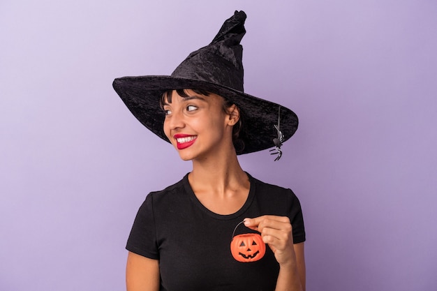 Young mixed race woman disguised as a witch isolated on purple background  looks aside smiling, cheerful and pleasant.