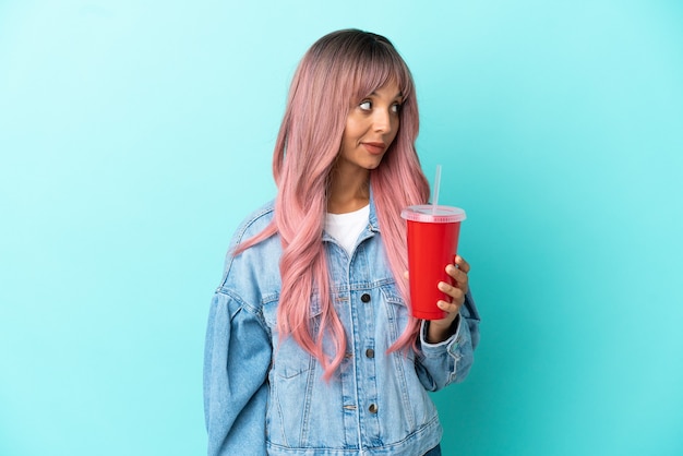 Young mixed race woman drinking a fresh drink isolated on blue background looking to the side