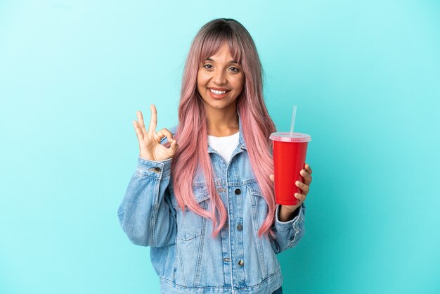 Young mixed race woman drinking a fresh drink isolated on blue background showing ok sign with fingers