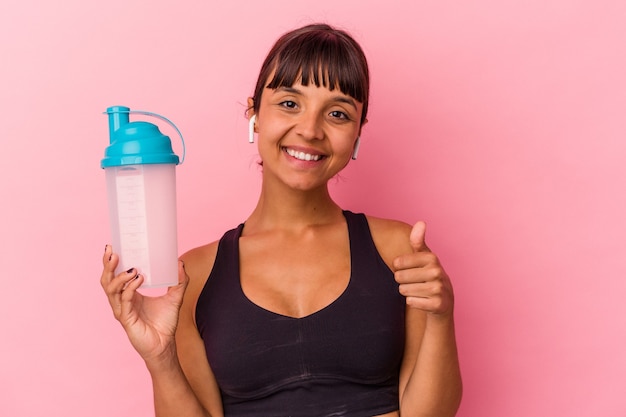 Young mixed race woman drinking protein shake isolated on pink background smiling and raising thumb up