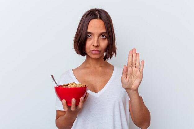 Young mixed race woman eating cereals isolated on white wall standing with outstretched hand showing stop sign, preventing you.