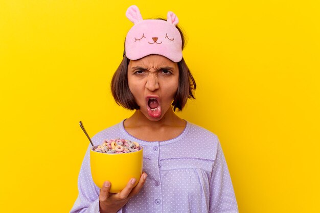 Молодая женщина смешанной расы ест хлопья в пижаме, изолированной на желтой стене, кричит очень сердито и агрессивно.