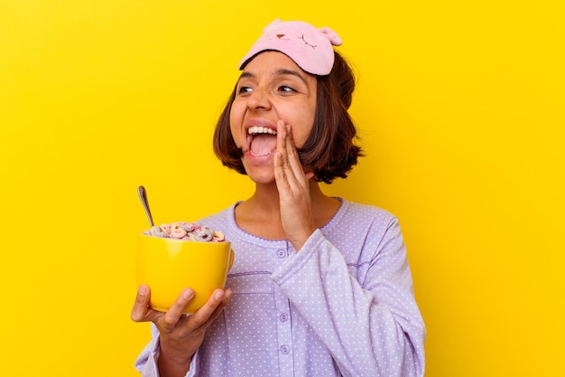 Young mixed race woman eating cereals wearing a pijama isolated on yellow wall shouting and holding palm near opened mouth.