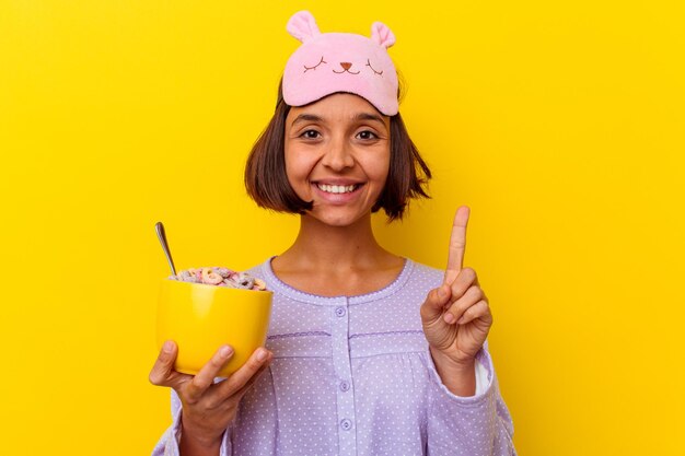 Young mixed race woman eating cereals wearing a pijama isolated on yellow wall showing number one with finger.