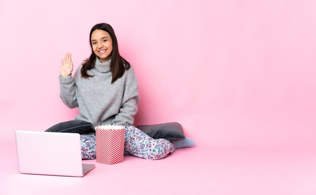 幸せな表情で手で敬礼ラップトップで映画を見ながらポップコーンを食べる若い混血の女性