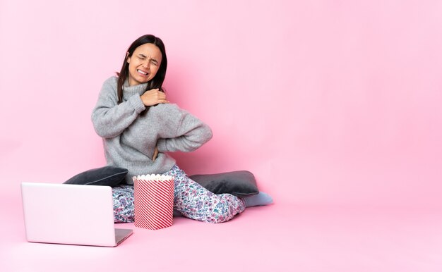 Giovane donna di razza mista che mangia popcorn mentre guarda un film sul laptop che soffre di dolore alla spalla per aver fatto uno sforzo